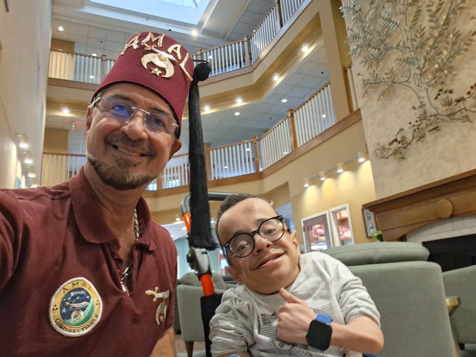 Nobre do AMAL Shriners com camisa vinho e fez cerimonial abraçado a criança sorridente no lobby de um hospital Shriners — momento de proximidade que ilustra o cuidado humanizado da fraternidade