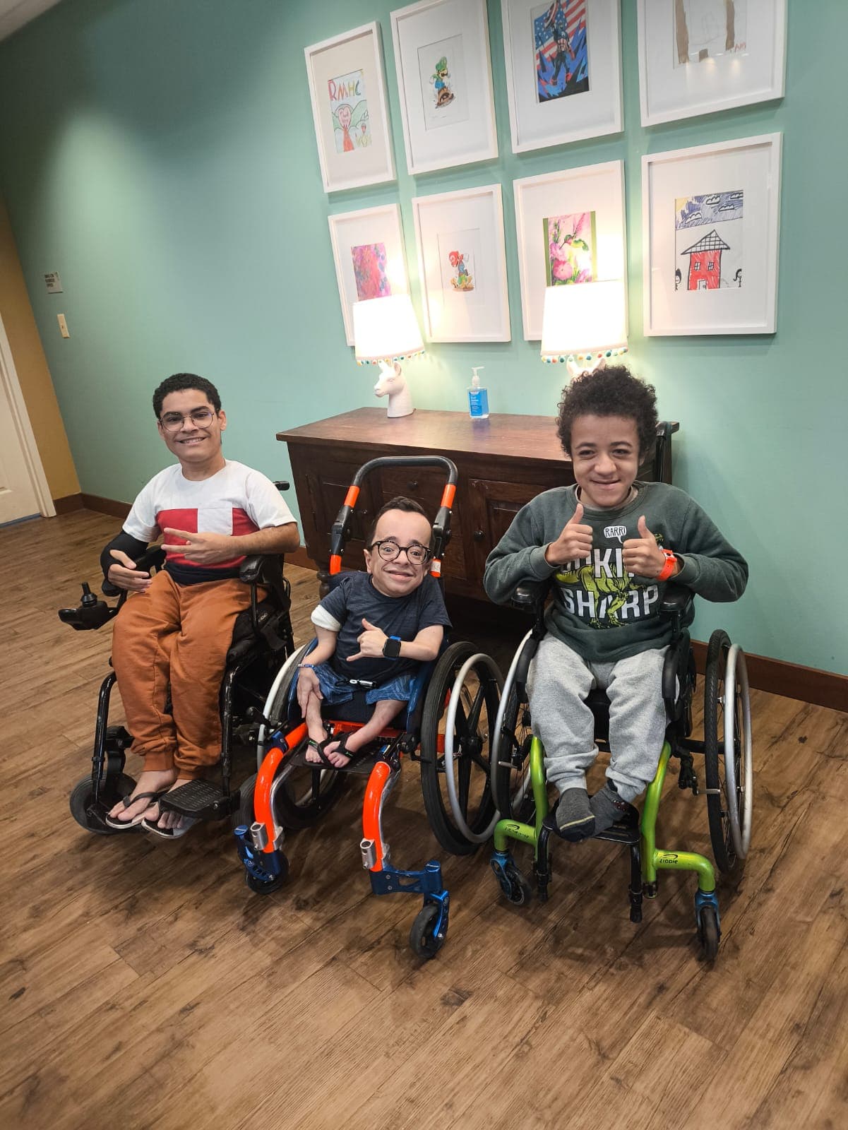 Três crianças em cadeira de rodas — Luiz Miguel, Francisco e Raphael — sorrindo e fazendo gestos de positivo em unidade da Ronald McDonald House Charities em Chicago, pacientes acompanhados pelos Shriners