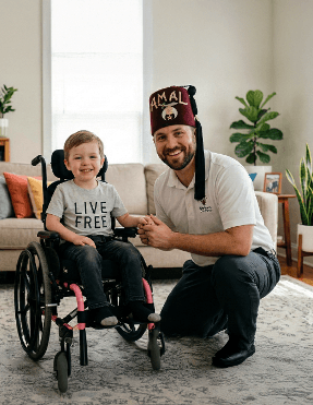 Voluntário Shriner sorridente ao lado de criança em cadeira de rodas com camiseta 'Live Free', em ambiente acolhedor de hospital pediátrico — retrata a proximidade humana que define o cuidado Shriners